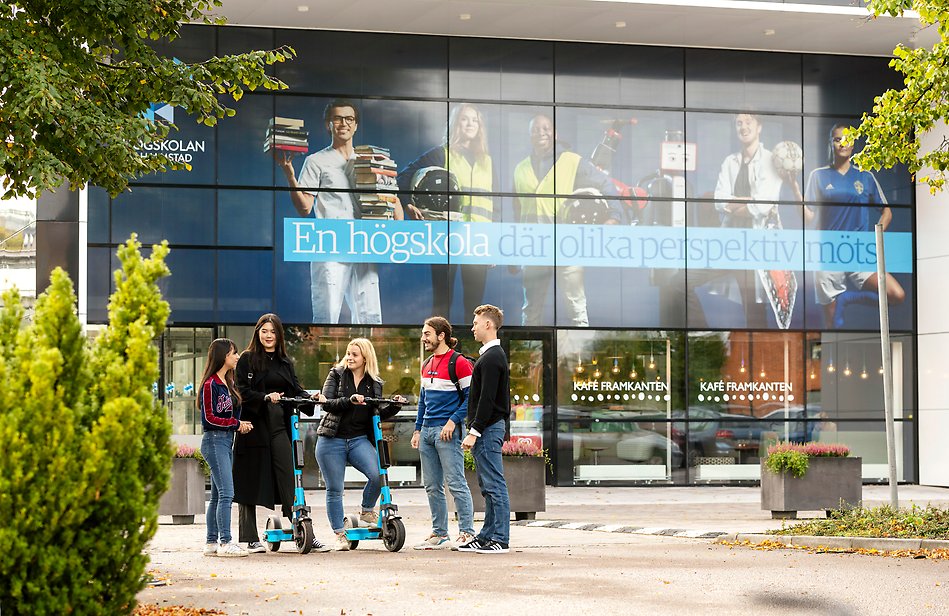 A group of students are conversating in front of the University entrance. Two of them are having electric scooters with them. Photo.
