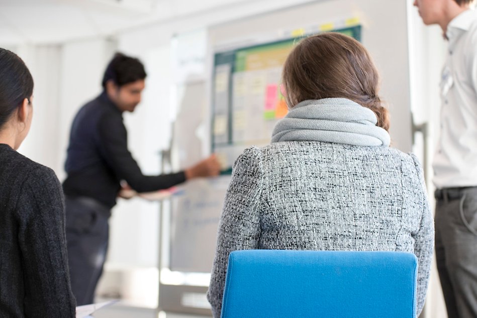 En person står vid en whiteboardtavla, tre andra personer tittar på tavlan. Foto.