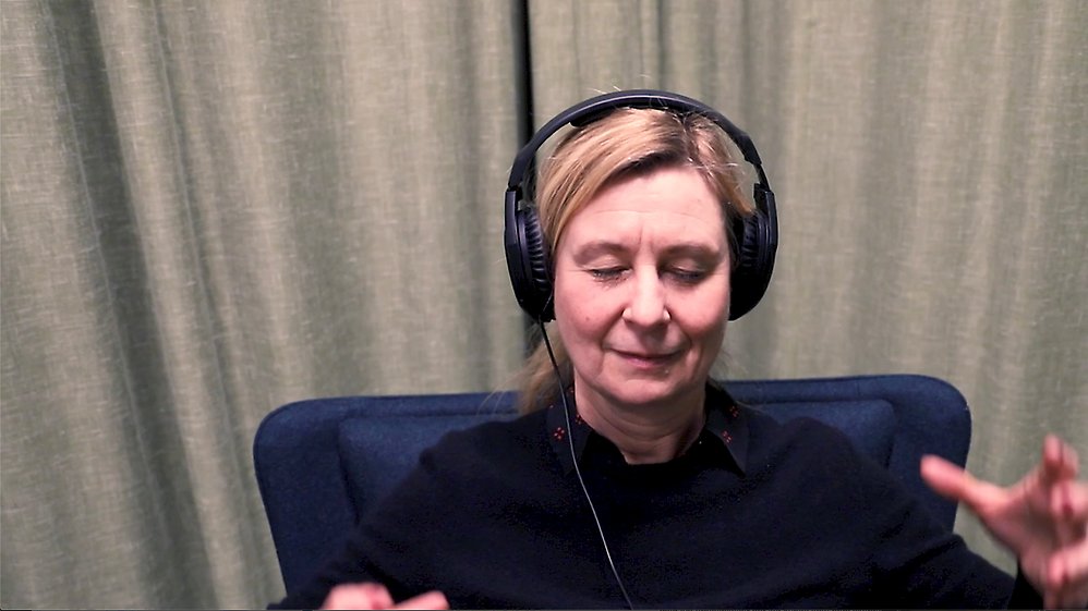 En blundande kvinna med hörlurar som sitter i en fåtölj. Foto.
