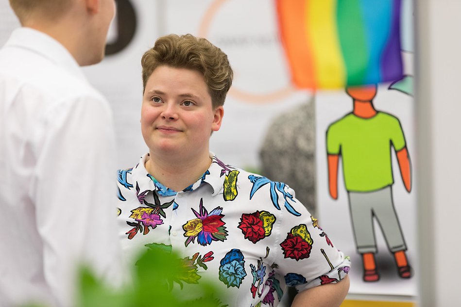 En person i färgglad skjorta pratar med någon. En prideflagga i bakgrunden. Foto.