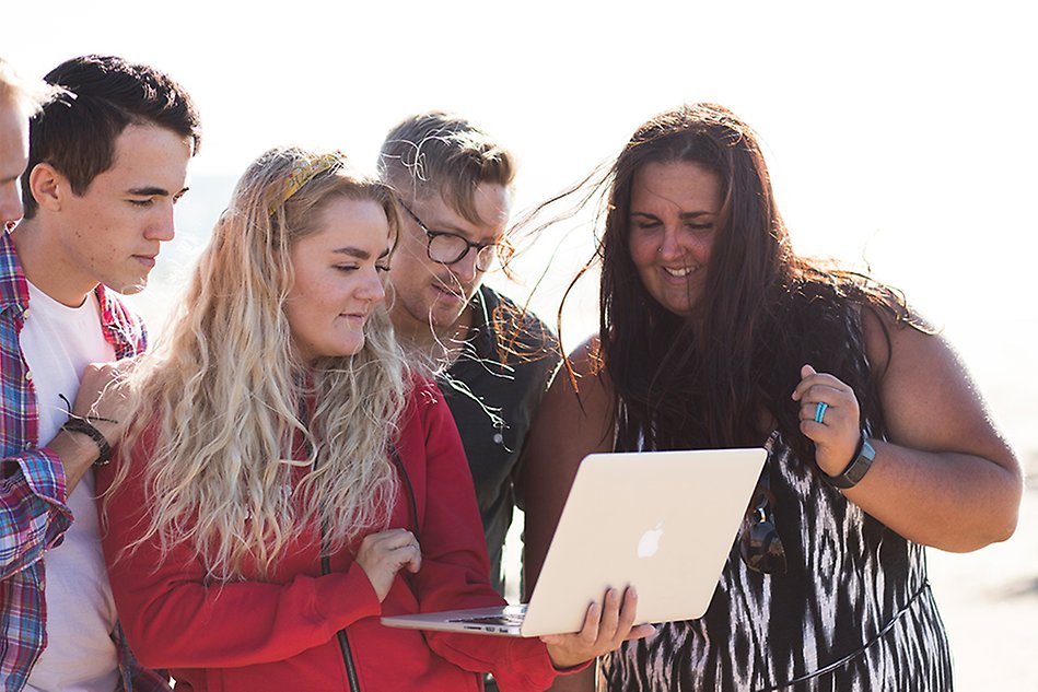 A group of people standing around a woman holding a laptop, looking at the screen. Photo.