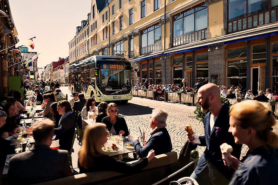 A bus is driving in a city. People are sitting outside at restaurants along the way. Photo.