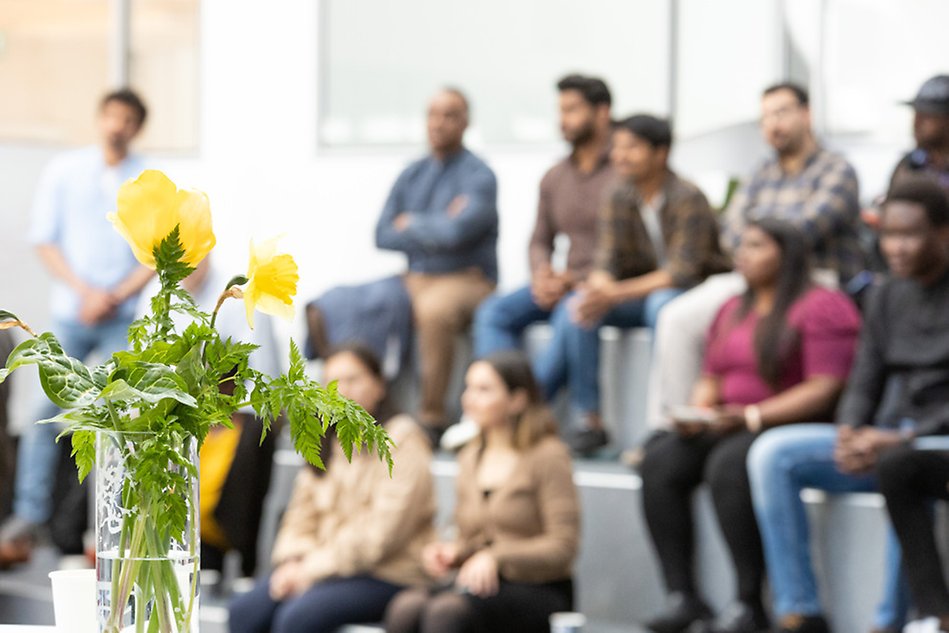 Folk i en publik syns i bakgrunden och en gul blomma är i fokus. Foto. 