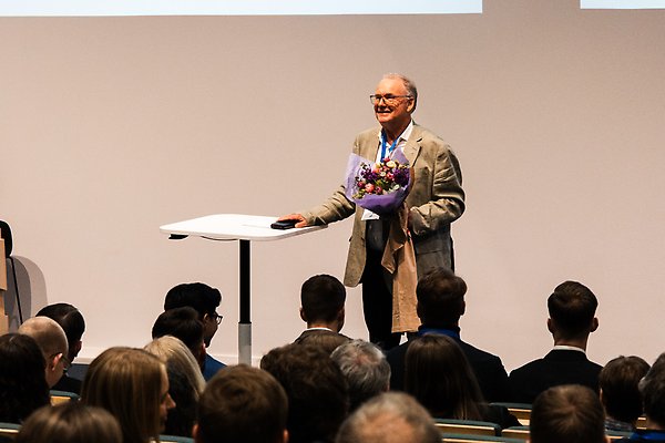 En man i kostym står på scen och ler mot publiken. Han håller i en bukett blommor. Foto.