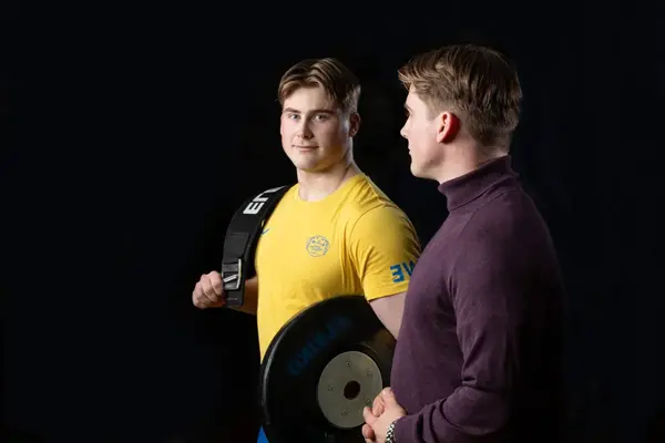 Två män står vända mot varandra. En är klädd i sportkläder med tyngdlyftningsvikter i händerna. Foto