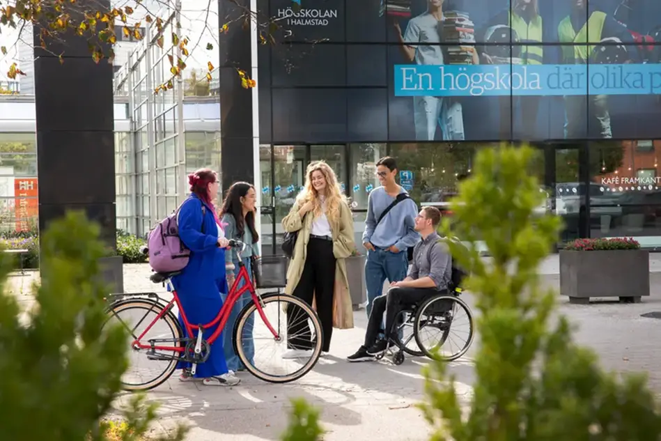 Fem unga personer i grupp står framför en byggnad utomhus i solen. En av personerna sitter i rullstol och en står vid en cykel. Foto.