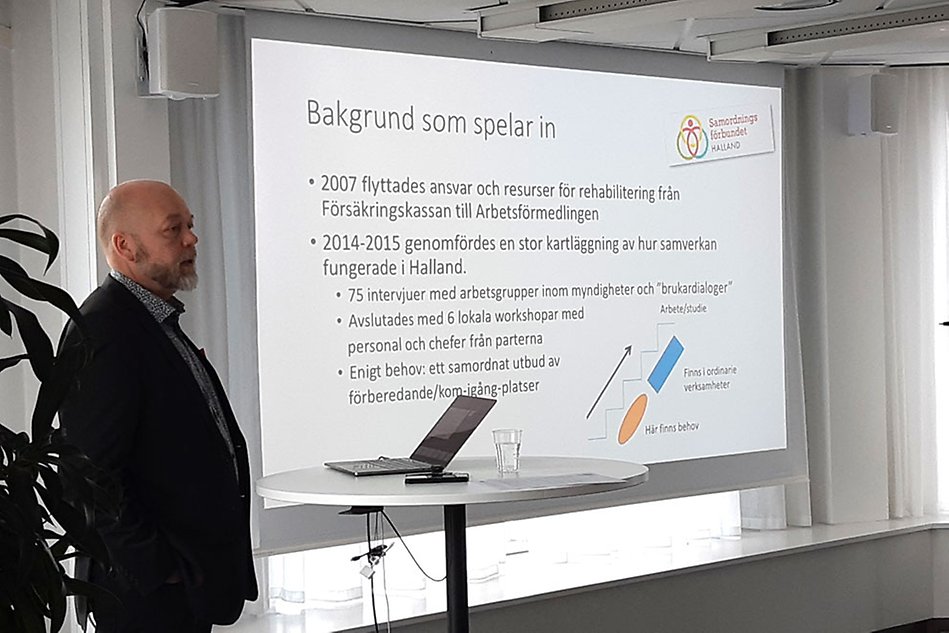 Man i kavaj och skägg föreläser vid storskärm. Foto.