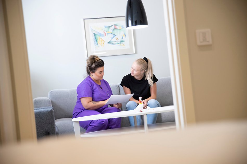En kvinna i lila sjuksköterskekläder pratar med en annan kvinna. De sitter i en soffa. Foto.