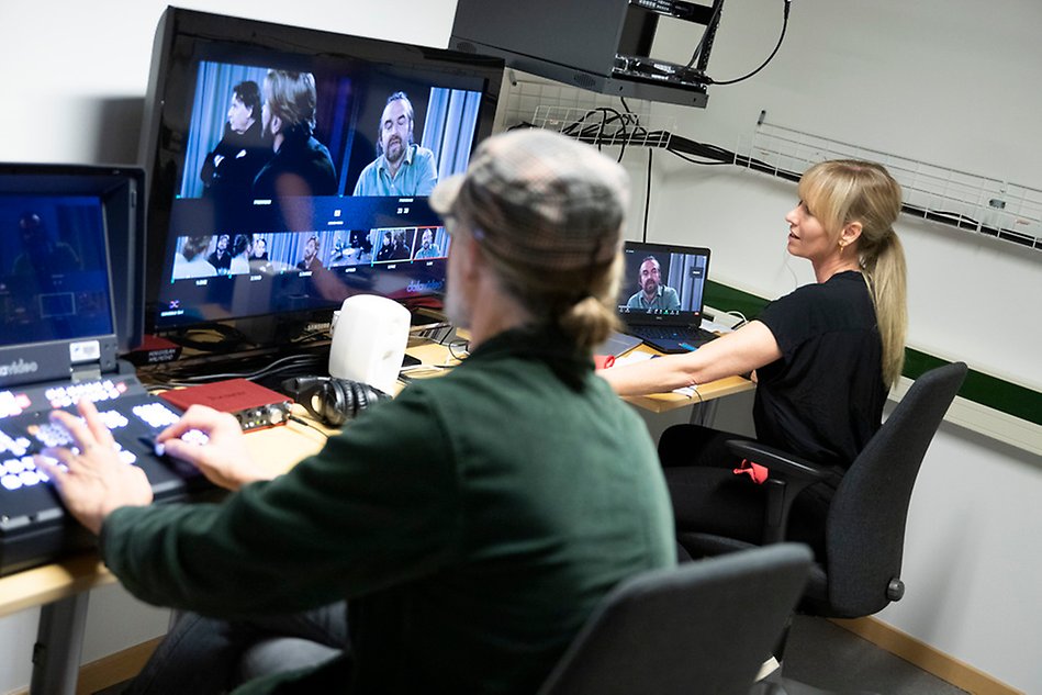 En man och en kvinna sitter i ett kontrollrum och tittar på en skärm som visar bilder från en tv-studio. Foto.