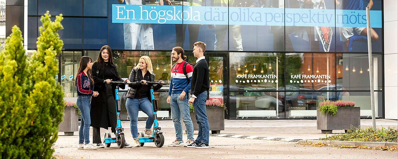 En grupp på fem unga personer står framför en byggnad med texten ”En högskola där olika perspektiv möts”. Två av personerna står på var sin elsparkcykel. Foto.