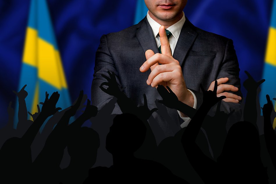 Man i kostym står med svenska flaggan bakom sig och en publik framför. Foto.