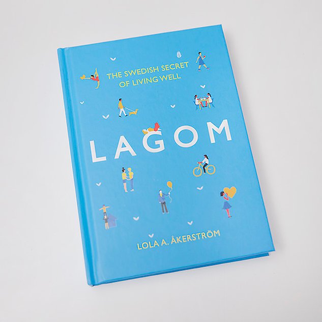 En ljusblå bok med små figurer och texten ”Lagom” ligger mot en vit bakgrund. Foto.