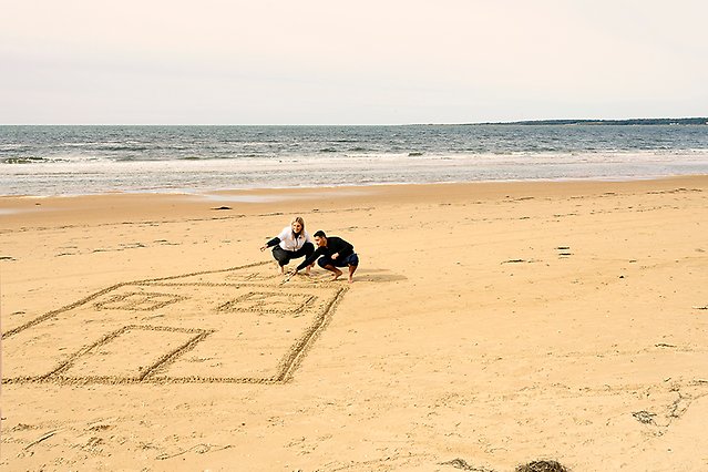 Två personer ritar ett stort hus i sanden på en strand. I bakgrunden syns havet. Foto.
