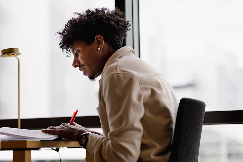 A person with black hair sitting at a table holding a pencil. The person is in profile view. Photo. 