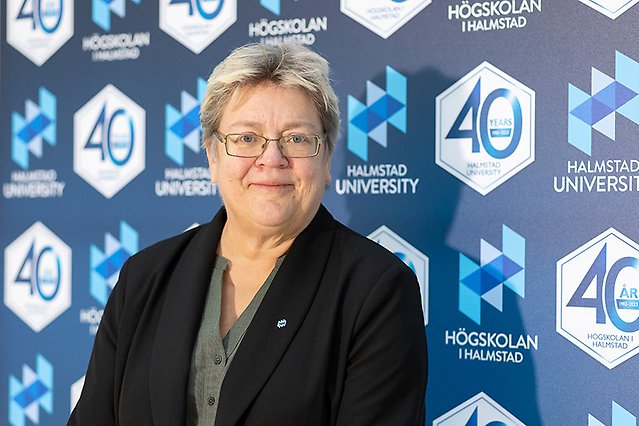 Blond, korthårig kvinna i svart kavaj och glasögon står framför en jubileumsvägg och ler mot kameran.