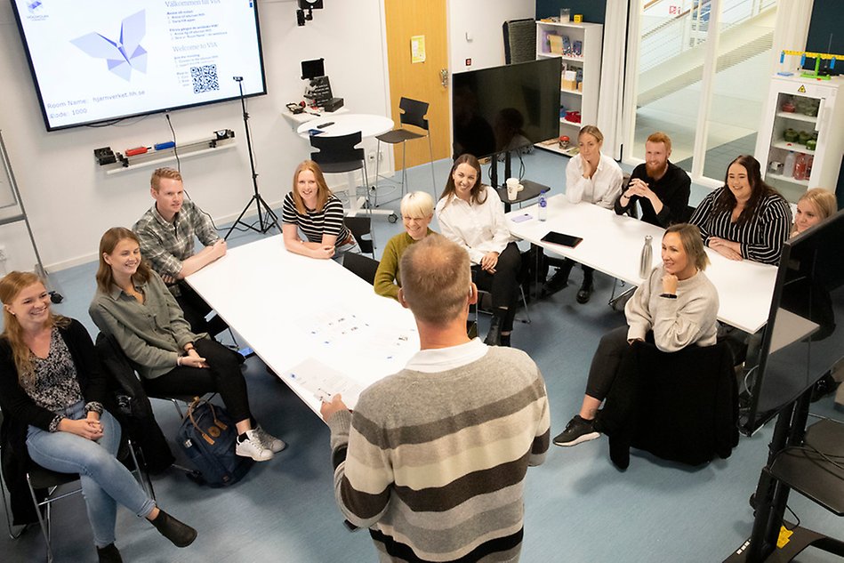 En person i beige-svart tröja står inför en grupp personer som sitter vid vita bord