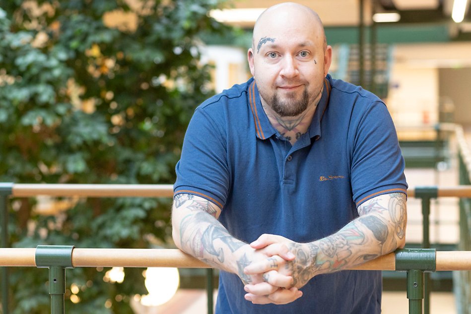 En man lutar sig mot ett träräcke och tittar in i kameran. I bakgrunden syns gröna växter. Foto.
