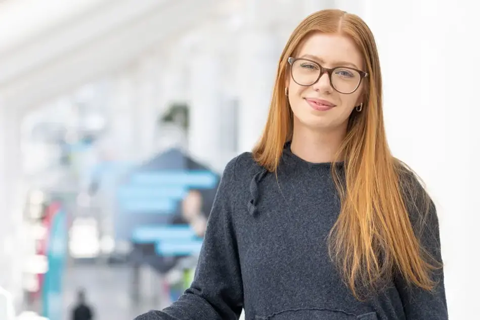 Student i rött långt hår, glasögon och hoodie står lutad mot en gradäng och ler mot kameran.