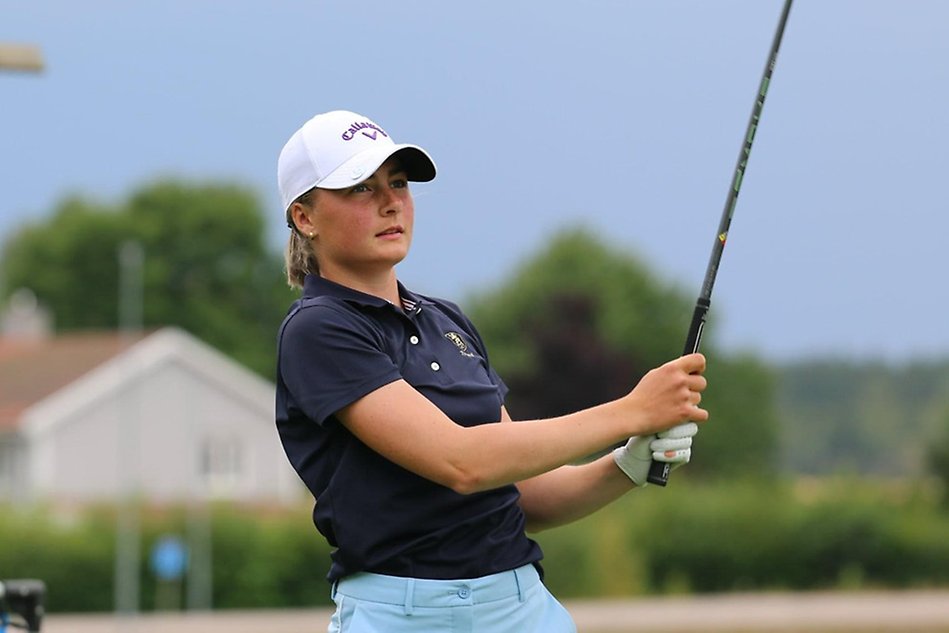 Tjej i vit keps och blå pikétröja slår ett golfslag utomhus. Foto.