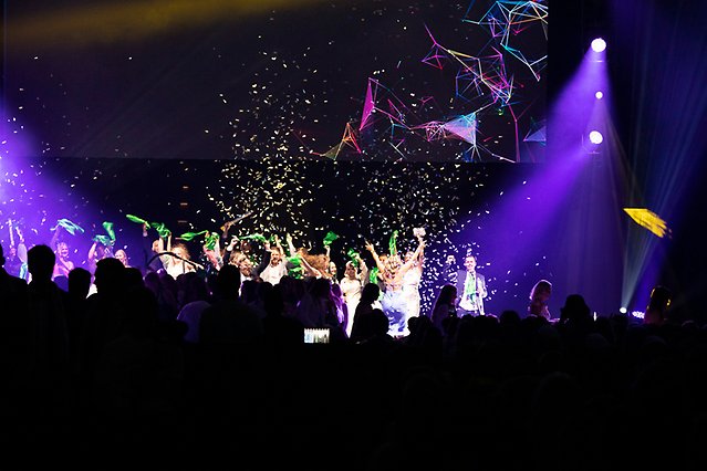 Unga människor står på en scen med viftande sjalar och under ett regn av konfetti. Framför scenen syns publik. Foto.
