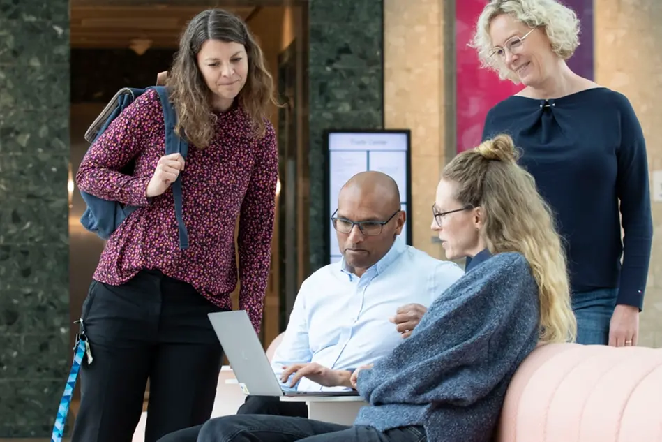 A group of people gathering around a pink couch looking at a computer screen. Photo.
