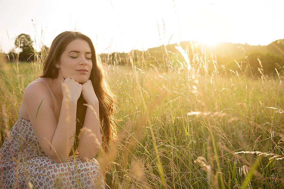 Woman with long brown hair, sitting on a meadow in sunlight with her eyes closed. Photo. 