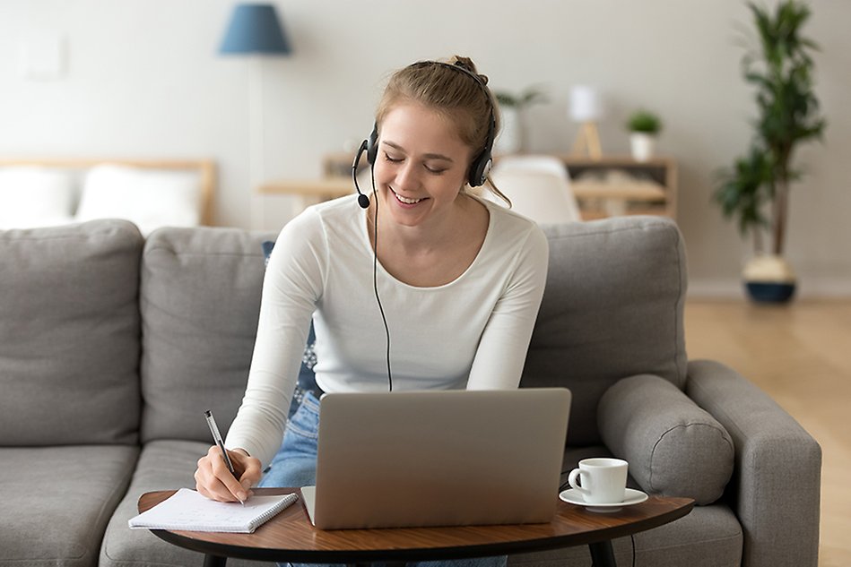 En leende kvinna med hörlurar i öronen sitter i en soffa i hemmiljö framför en dator och skriver i ett block som ligger på bordet. Foto.