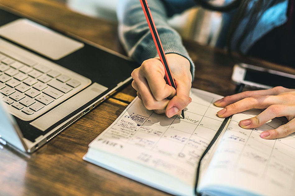 A hand is seen writing with a pencil in a calendar. To the left is a laptop. Photo.