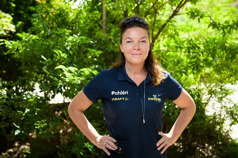 A middle-aged woman with brown hair is standing outside in a blue polo shirt. She smiles at the camera with her hands on her hips. In the background are bushes. Photo.