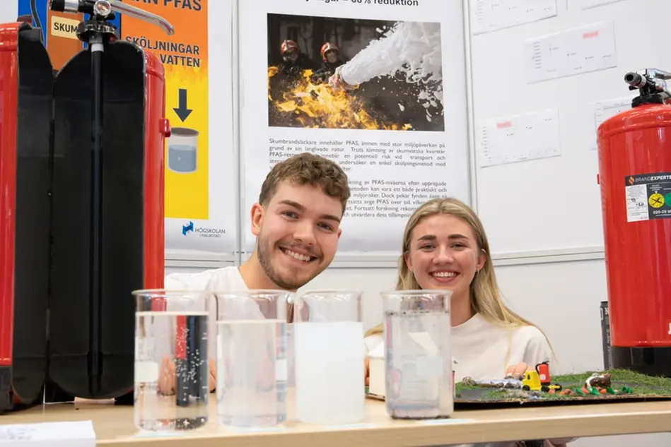 Två studenter i vit t-shirt hukar ner bakom ett bord och ler mot kameran. Framför dem finns brandsläckare och glas. Foto.