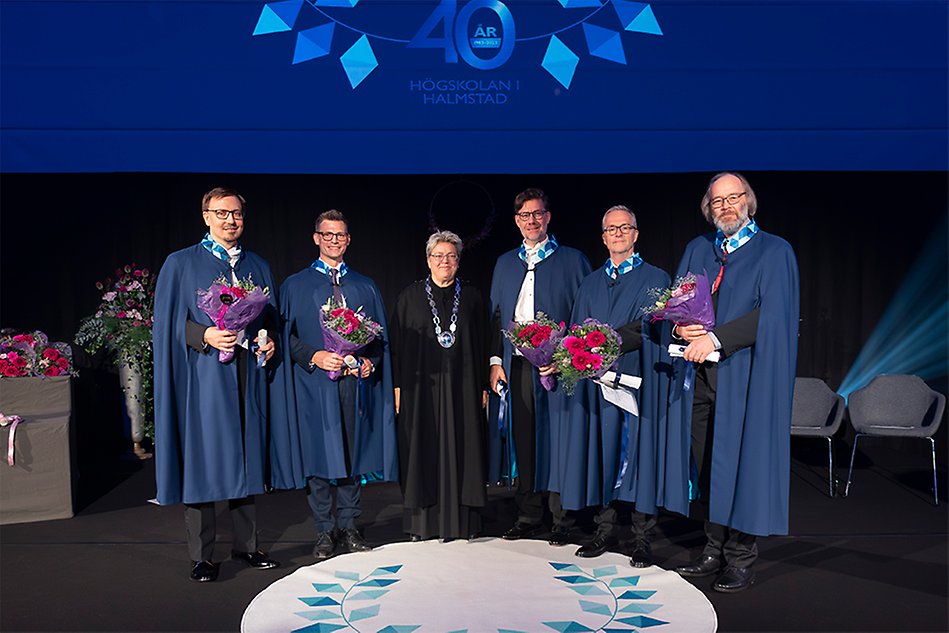 Five men in blue gowns and a woman in a black gown are lined up next to each other. The men have bouquets of flowers in their hands. Photo.