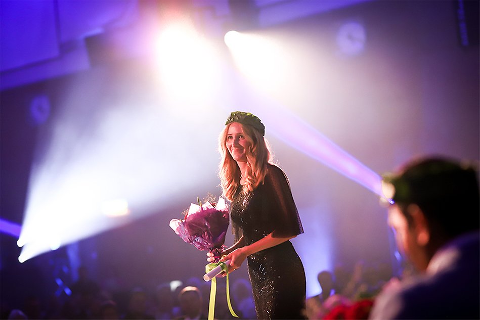 A woman with light long hair and a dark, glittering dress stands on a stage. In her hands she holds a bouquet of flowers and on her head she has a wreath. Photo.