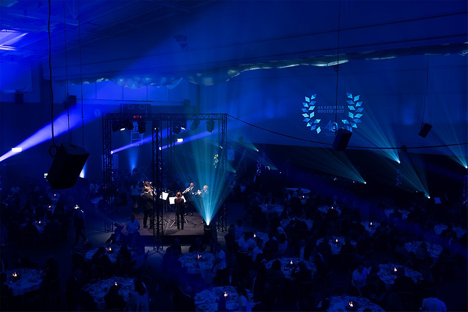 A dark room with round tables with white tablecloths and lit candles on the tables. The photo also shows a stage with a brass band. Photo.