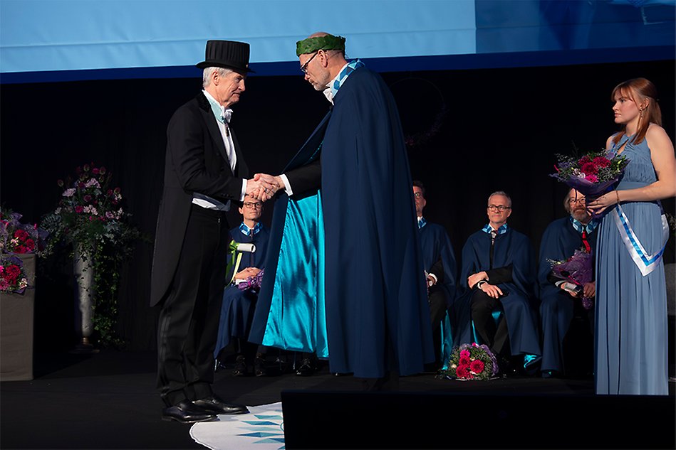 A man in a black gown and black hat stands on a stage and shakes hands with a man in a blue gown and a wreath on his head. Photo.