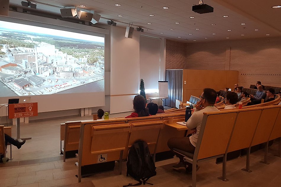 Bild av en föreläsningssal där människor tittar på en stor skärm som visar en industri.
