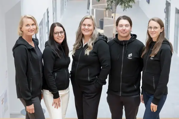 Five smiling people, dressed in identical black sweaters, stand in a row in a bright indoor setting. Photo.