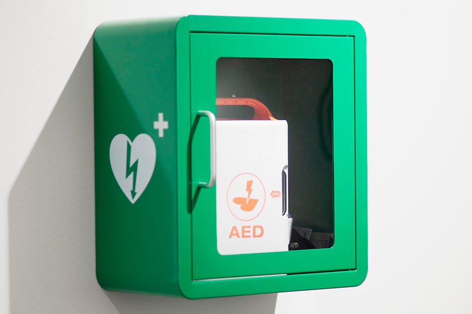 En grön låda fastmonterad på väggen. Genom en glasruta kan man se en röd hjärtstartarapparat. Foto. 