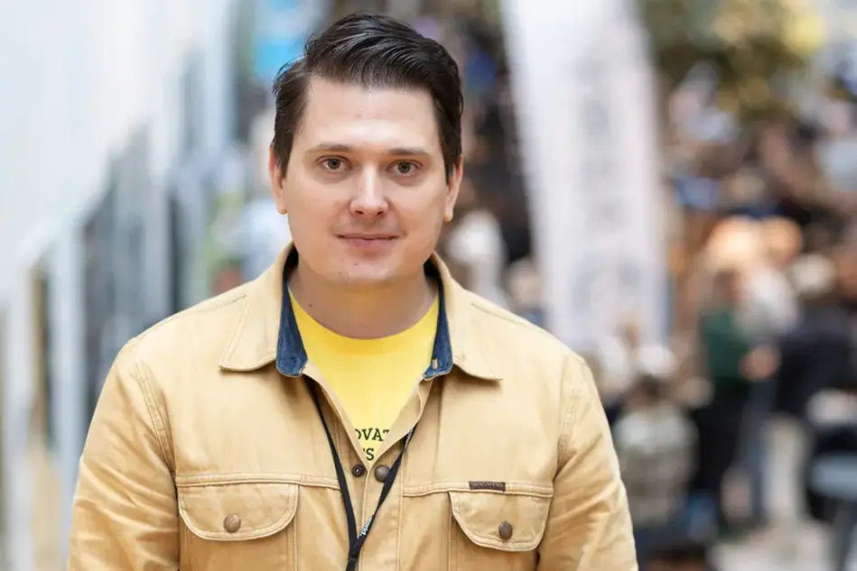 A brown-haired man wearing a beige shirt and a yellow T-shirt smiles at the camera. In the background, there is a crowd of people. Photo.