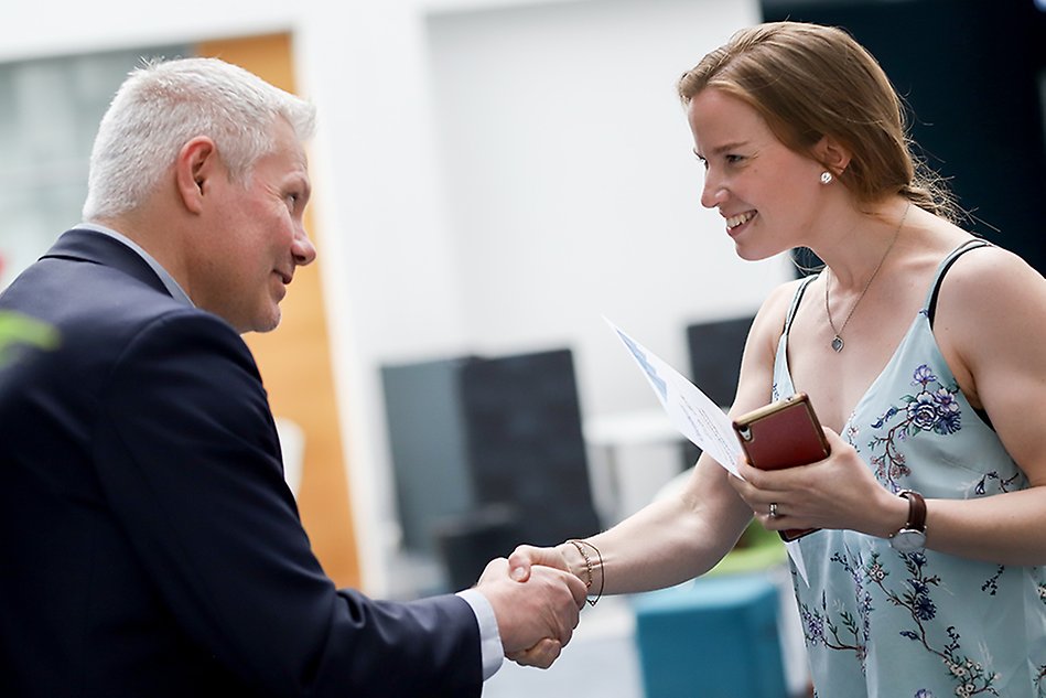 A man is handing over a certificate to a woman. There are shaking hands and smiling. Photo.