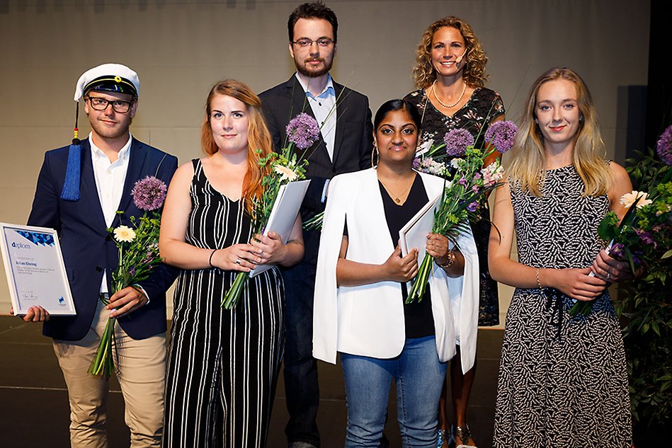 Six well-dressed people stand next to each other, all but one holding their diplomas and flowers. Photo.