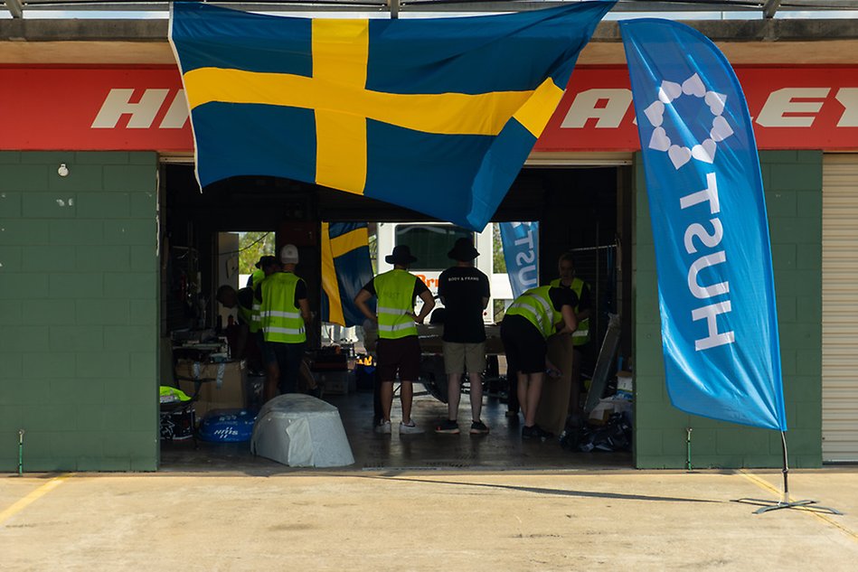 Personer som står i ett garage med ryggen mot oss. En stor svensk flagga och en flagga där det står HUST. Foto.