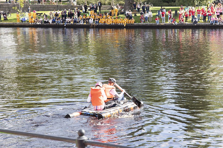 Two young men are sitting on a small raft in a river in an urban environment. They are wearing life jackets. Photo.