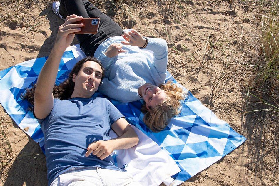 Två unga personer ligger på en handduk på stranden. Båda tittar in i en mobil och ler. Foto.