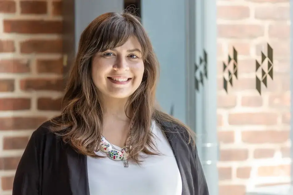 A woman in brown hair, white shirt and black cardigan stands in front of a brick wall and smiles to the camera.
