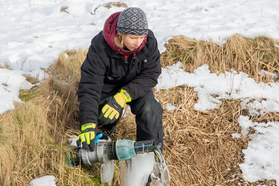 A person in a snowy landscape, holding their hand on a pipe. Photograph.