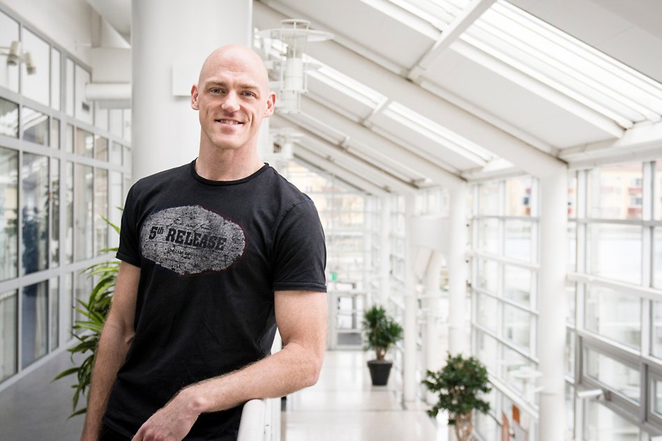 Man wearing a black t shirt smiling leaning against the railing of a balcony overlooking a corridor with glass walls. Photo.