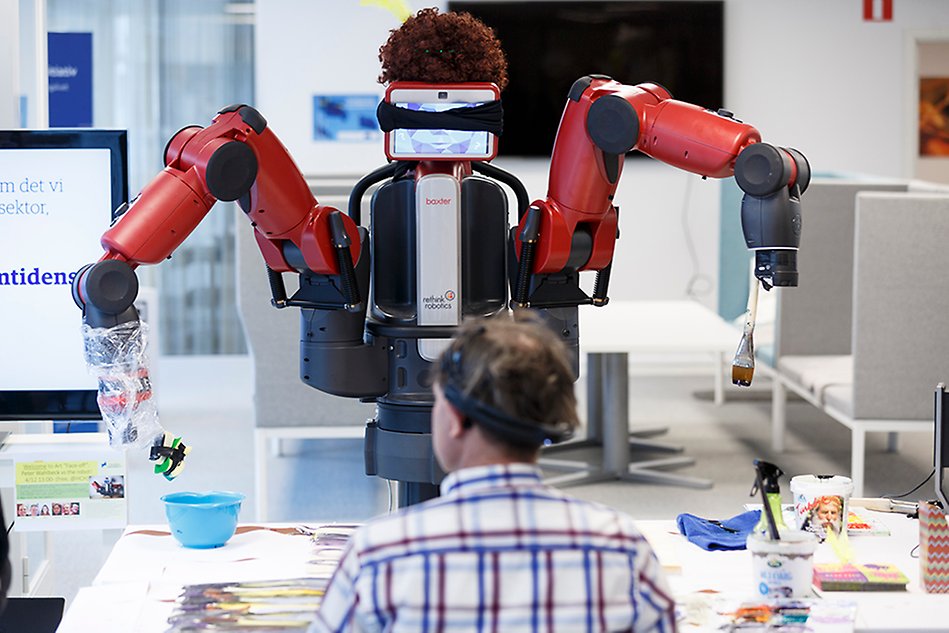 Red robot torso with arms, painting with brushes on paper on a table in front of it, a man is sitting across from the robot