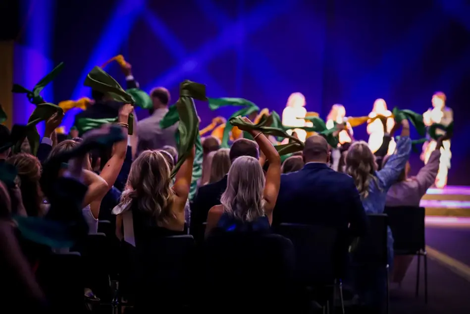 An audience of young and well-dressed people look up at a stage while waving colorful shawls. Photo.