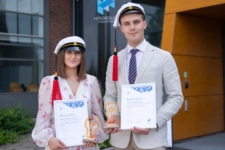 Två finklädda studenter håller i ett varsitt diplom och statyett och ler mot kameran. I bakgrunden finns en byggnad, buskage och en pelare med Högskolans logga på. Foto.