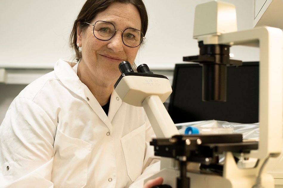 Woman in white lab coat looking into the camera. She sits by a microscope. Photo.